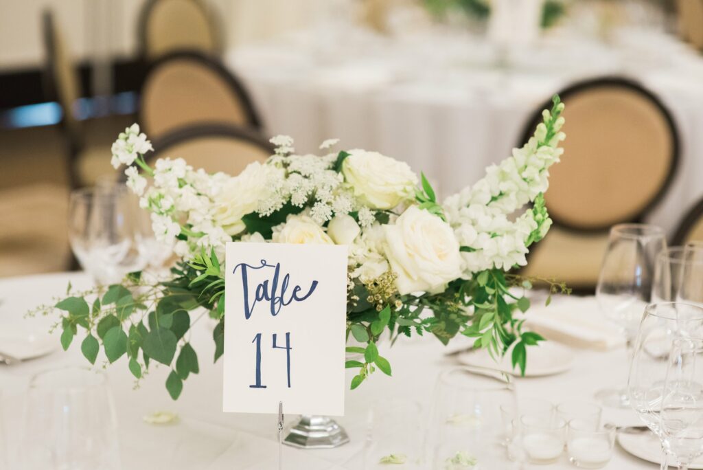 A floral centerpiece with white flowers and greenery sits on a round table with a card indicating Table 14.