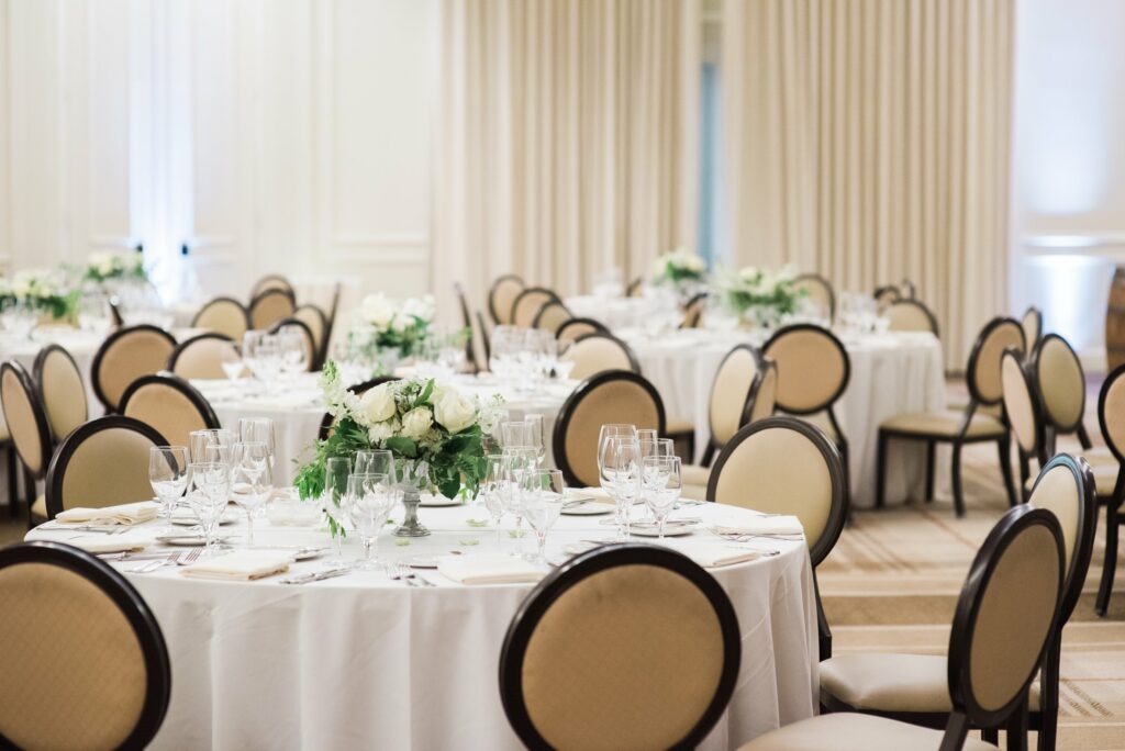A banquet hall is set up for an event with round tables, elegant place settings, and floral centerpieces. The chairs have beige seats and backs, and the room is illuminated with soft lighting.