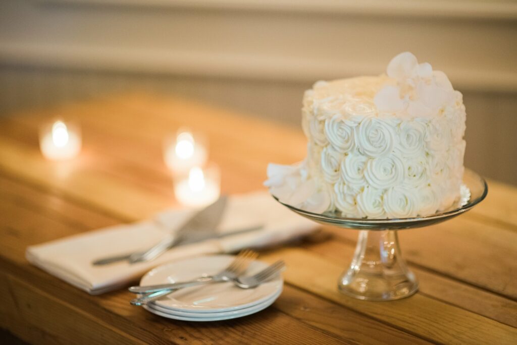 A white frosted cake with rose-like decorations on a glass stand, accompanied by plates, forks, and candles on a wooden table.