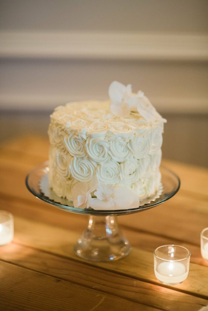 A white frosted cake with floral details sits on a glass pedestal, surrounded by small lit candles on a wooden surface.