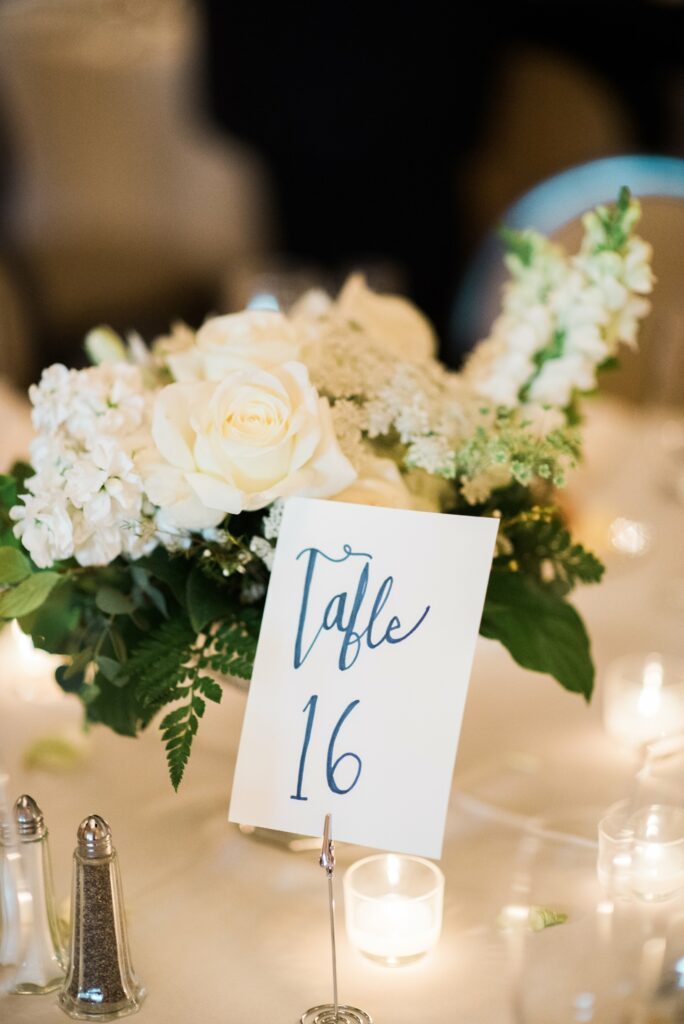 Table 16 centerpiece with white flowers, greenery, and a handwritten table number card. Surrounding the arrangement are small lit candles and salt and pepper shakers.