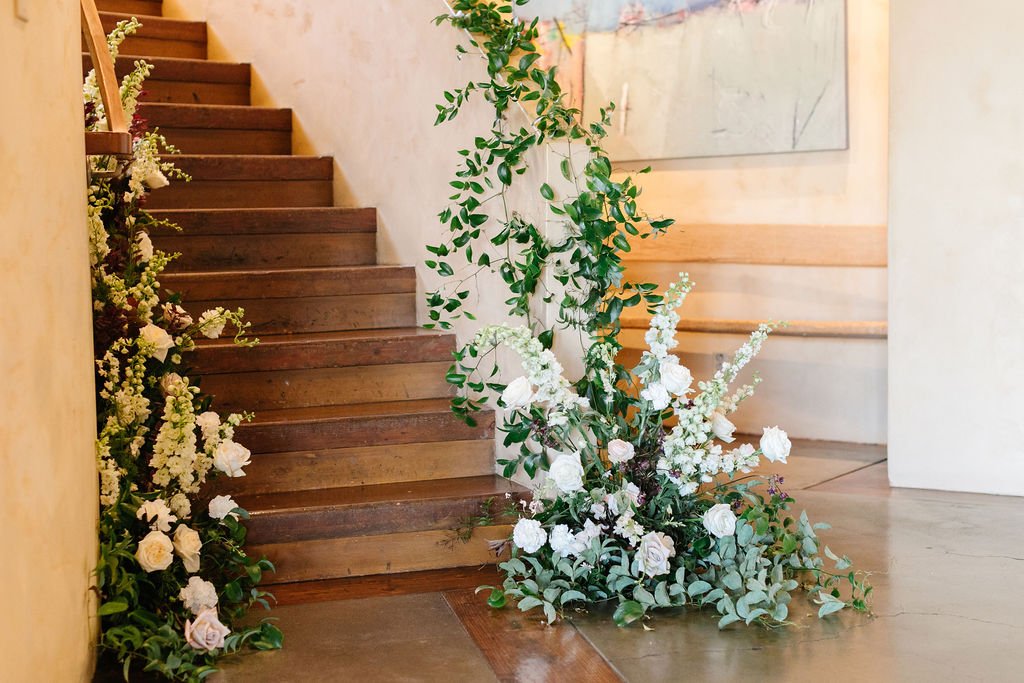 A wooden staircase decorated with white flowers and green foliage on the steps and at the base, beside a wall-mounted wooden bench.