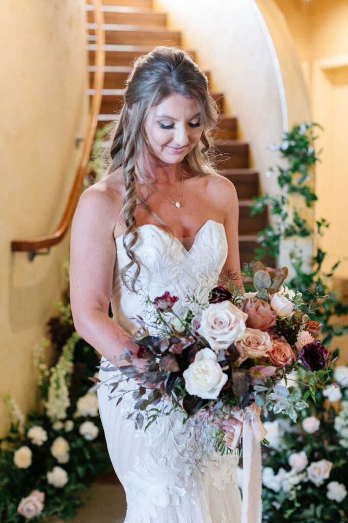 A bride in a white strapless wedding dress holds a bouquet of assorted flowers, standing in front of a staircase adorned with greenery.