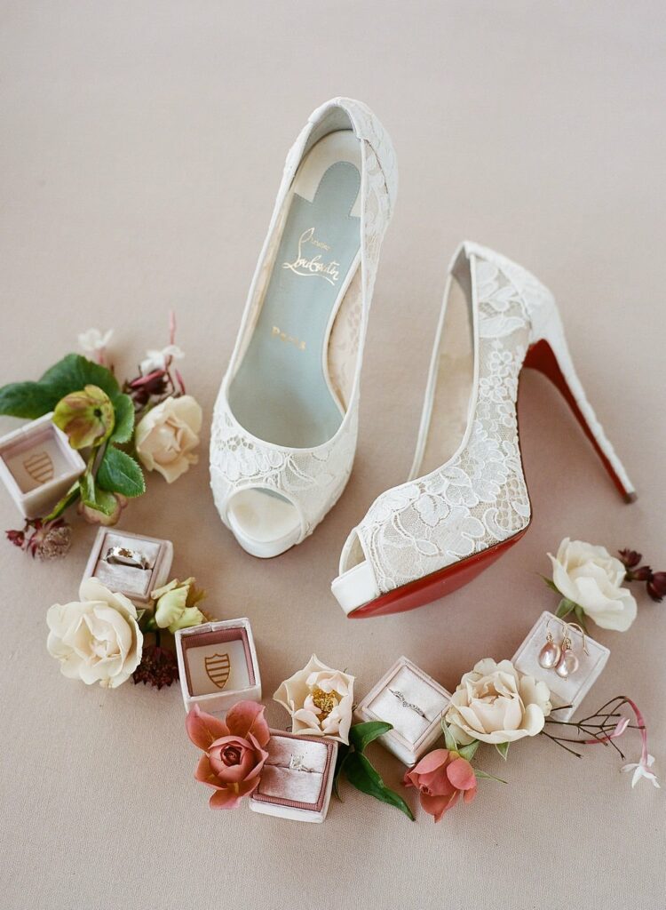 A pair of white lace high-heeled shoes with red soles, surrounded by various flowers and open jewelry boxes displaying rings and earrings.
