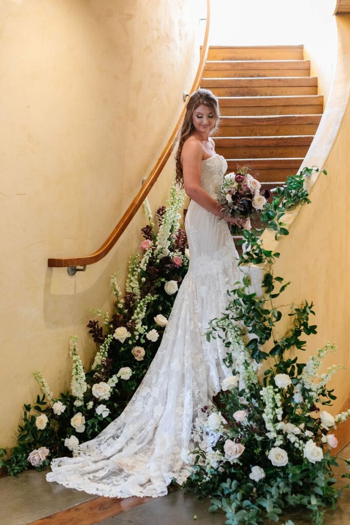 Bride in a white wedding dress stands on a staircase adorned with white and pink flowers, holding a bouquet and looking over her shoulder.