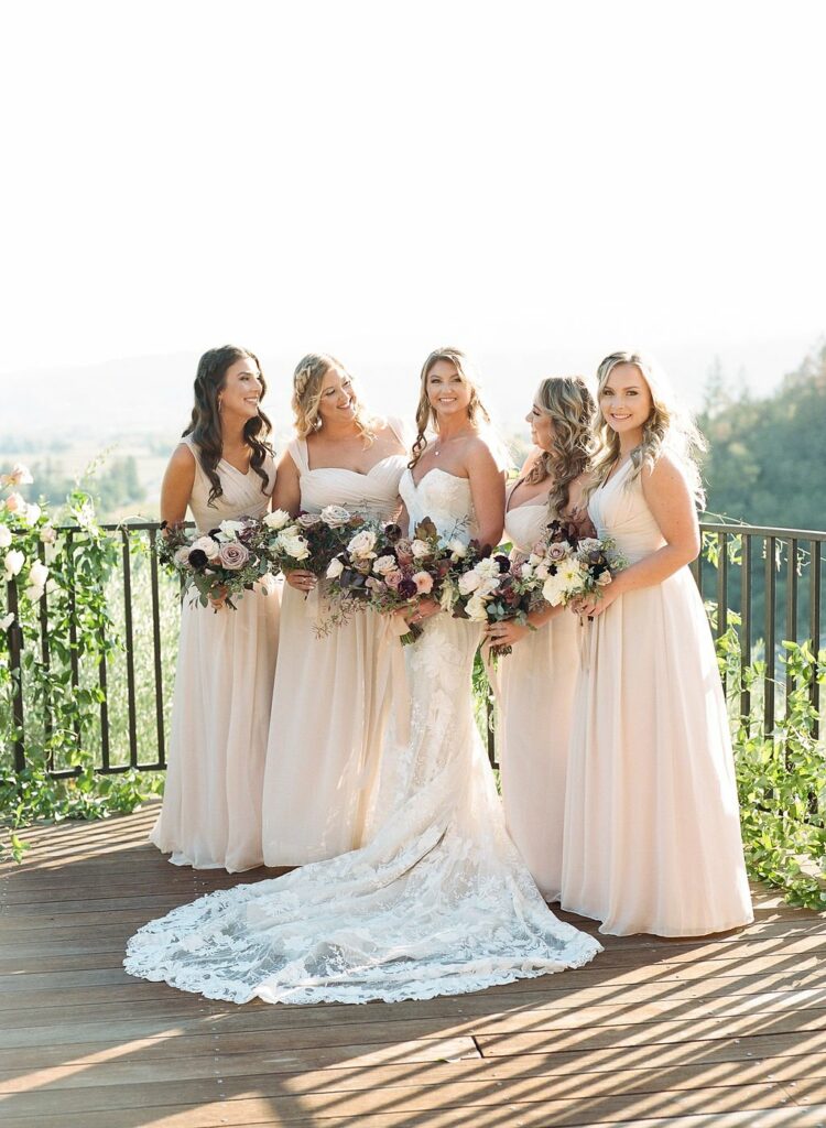 Five women in light-colored dresses, with the central bride in a white gown, pose together holding floral bouquets on a sunlit outdoor patio with greenery and a scenic background.