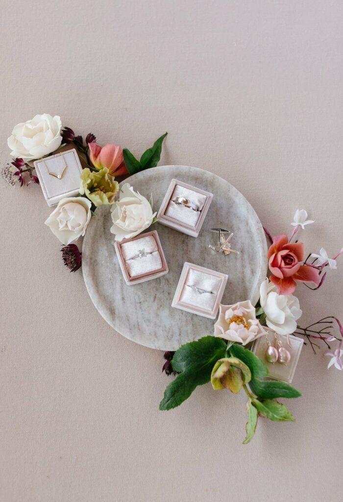 A round tray with four jewelry boxes containing rings, surrounded by white and pink flowers on a beige background.
