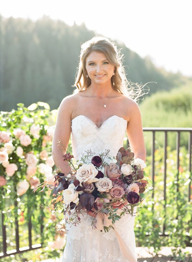 A bride in a strapless white wedding dress holds a bouquet of flowers outdoors with a greenery-filled background.
