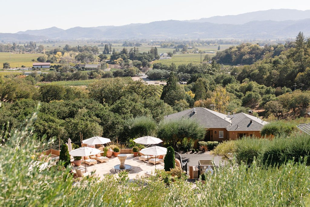 A scenic view of a countryside estate featuring several houses with gray roofs, surrounded by greenery and hills in the background. A patio area with umbrellas is visible in the foreground.