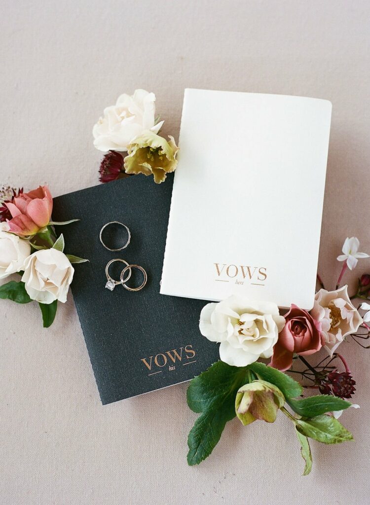 Two vow booklets, one black labeled "Vows his" and one white labeled "Vows hers," are surrounded by a variety of flowers and three rings on a light surface.