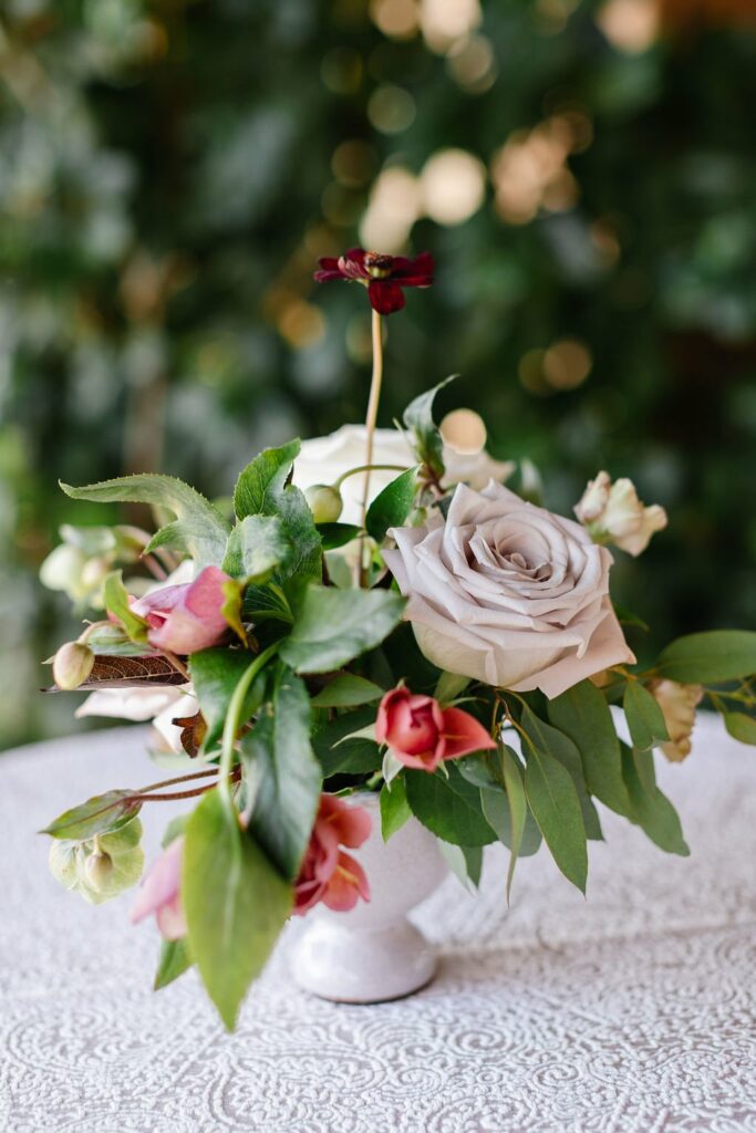 A floral arrangement featuring a white rose, pink flowers, and green leaves in a white vase, placed on a lace-covered surface.