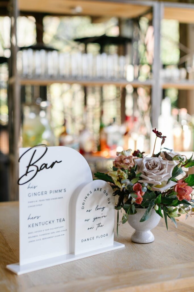 Bar menu featuring Ginger Pimm's and Kentucky Tea next to a floral arrangement on a wooden table with glass shelves and bottles in the background.
