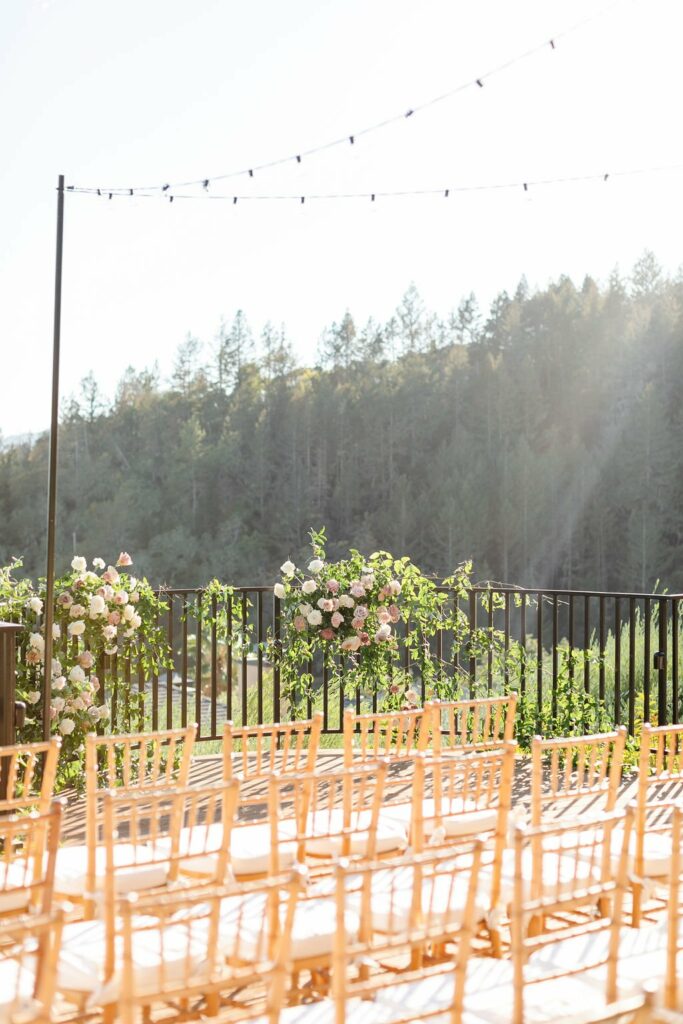 Outdoor wedding ceremony setup with rows of wooden chairs and floral arrangements on both sides of the aisle. A string of lights is hung above the seating area with a forested backdrop in the distance.