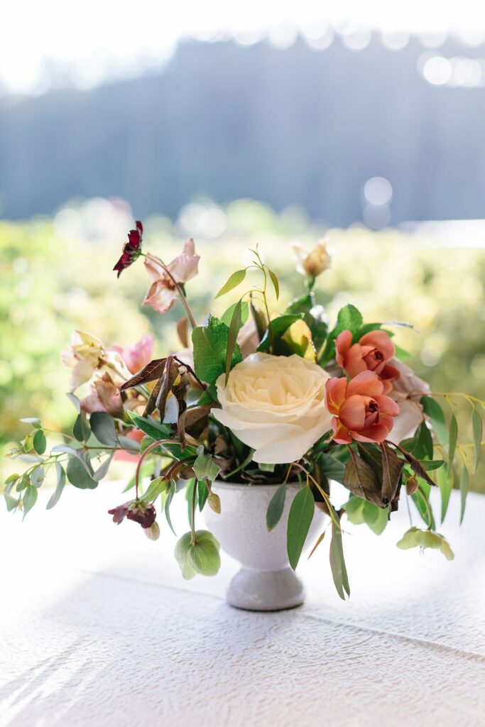 A white vase holds a bouquet of flowers, including a large white rose, pink blooms, green leaves, and branches, placed on a table with a blurred outdoor background.