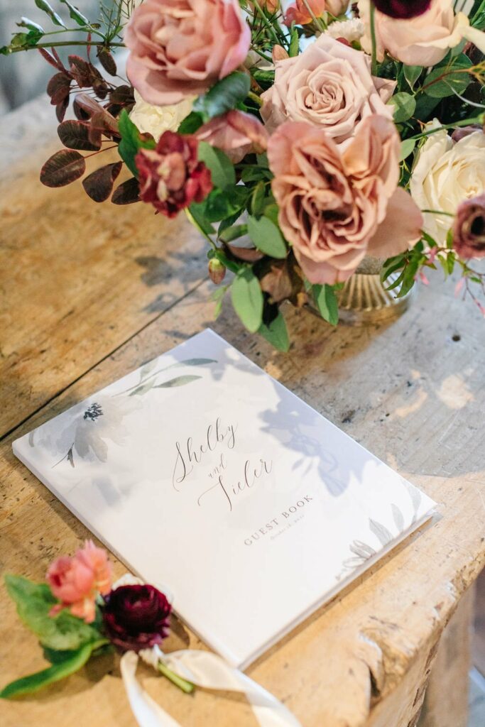 A guest book with "Shelby and Tyler" written on the cover is placed on a wooden table next to a floral arrangement with pink and red roses.
