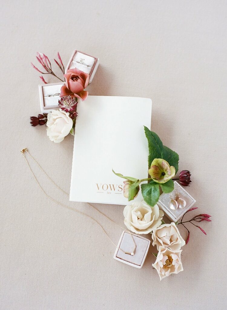A wedding vows booklet surrounded by flowers, jewelry boxes with rings, and a necklace with a pendant.