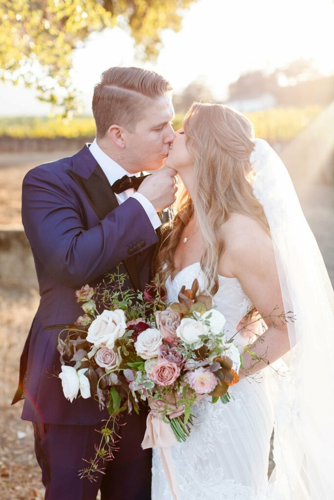 A bride and groom, in wedding attire, share a kiss outdoors while holding a bouquet of flowers.