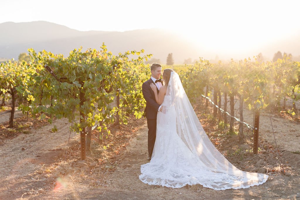 A bride and groom stand closely together in a vineyard at sunset. The bride wears a long white gown with a veil, while the groom is in a dark suit with a bow tie.