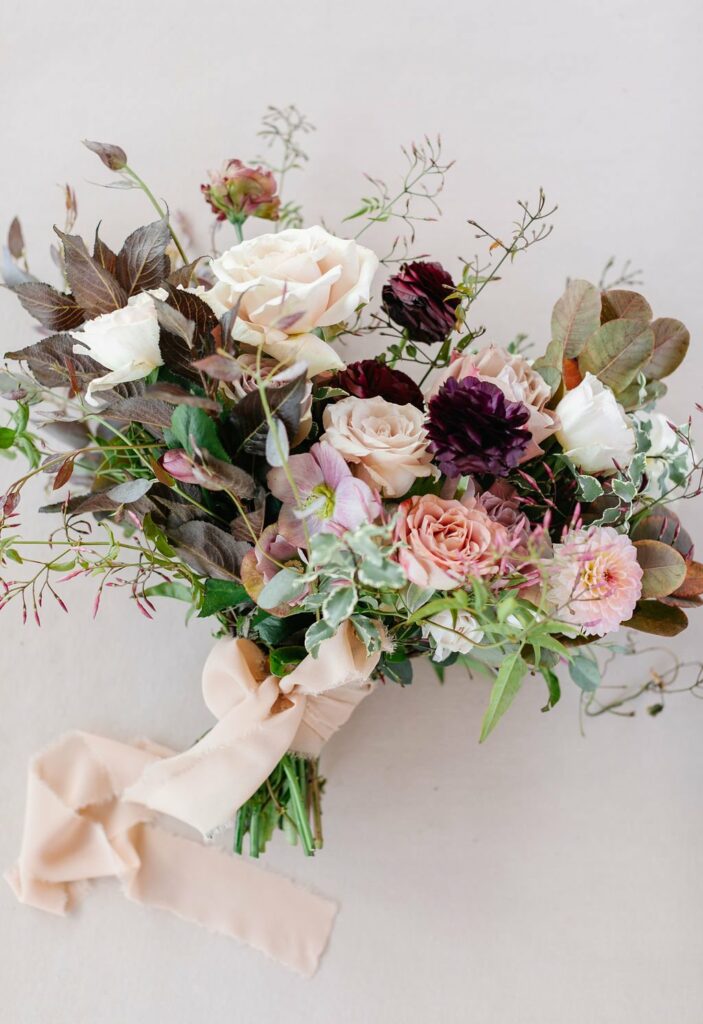 A bouquet of flowers featuring roses and various leaves and greenery, tied together with a beige ribbon.
