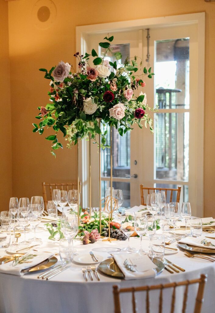 A round table set for a formal event with a tall floral centerpiece, white linens, glassware, and gold cutlery, situated in a room with double glass doors in the background.