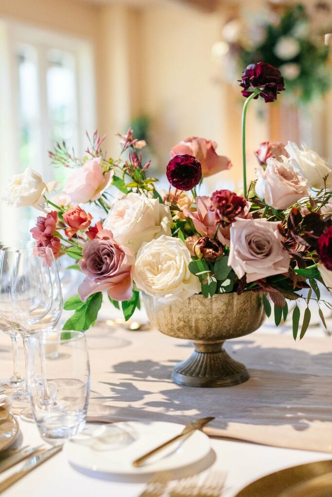 Elegant centerpiece featuring a mix of white, blush, and burgundy flowers in a gold bowl, surrounded by empty wine glasses and cutlery on a neatly set dining table.