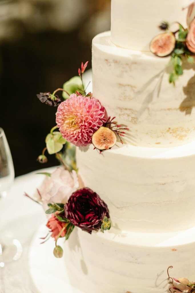 Close-up of a three-tiered white wedding cake adorned with assorted flowers, including pink dahlias and red blooms, and sliced figs. A wine glass is visible on the table beside the cake.