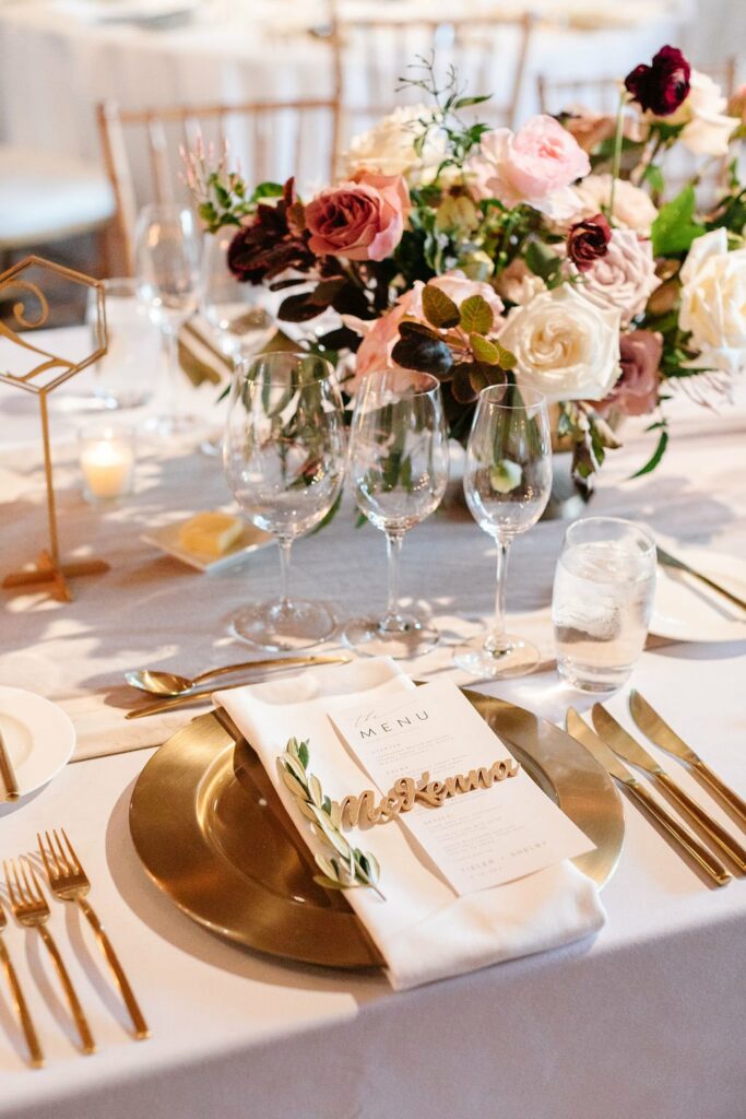 Elegant table setting with a gold charger, cutlery, glassware, floral centerpiece, and personalized name card beside a menu.