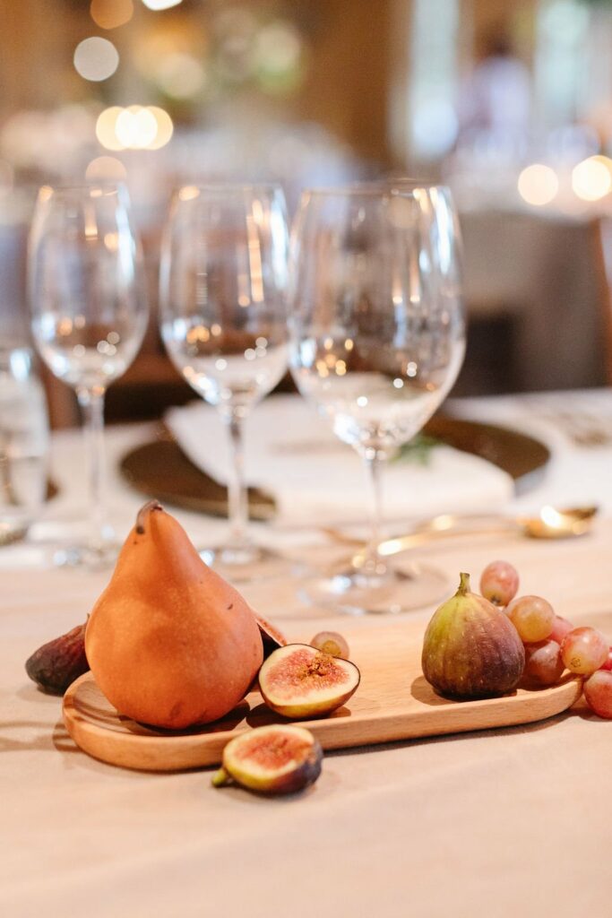 A table set with empty wine glasses and a wooden board featuring a pear, halved figs, a whole fig, and a small bunch of grapes.