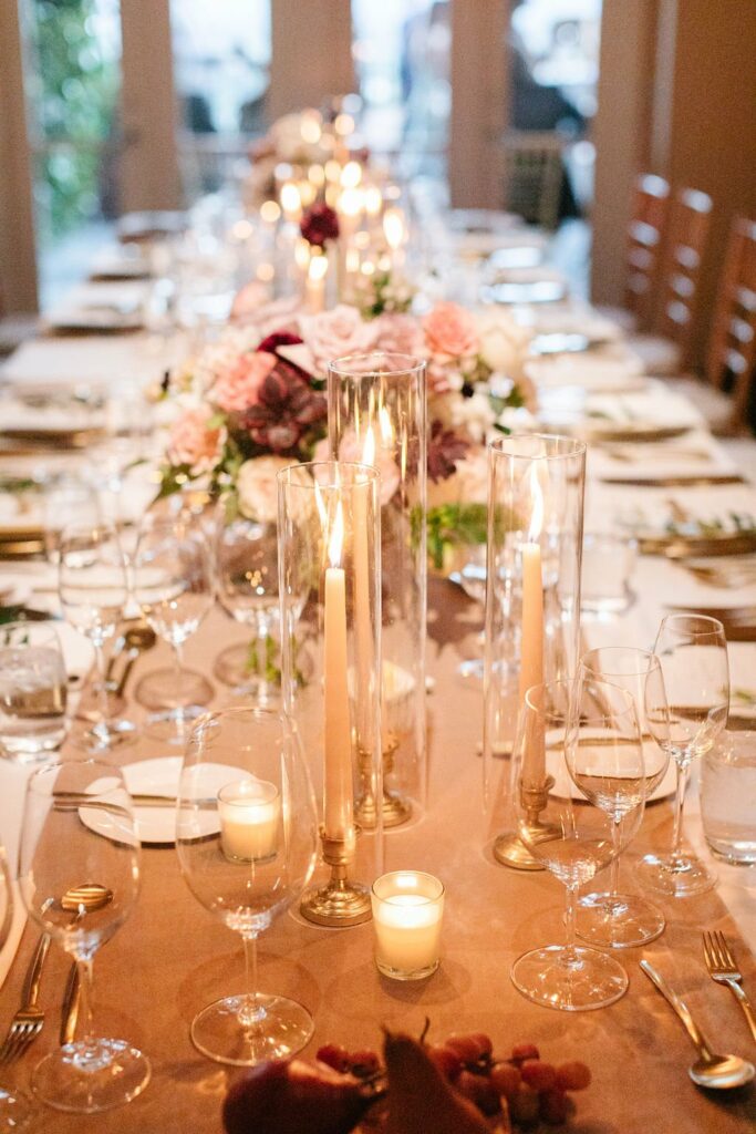 A long dining table is decorated with lit candles, floral arrangements, and set with plates, glasses, and silverware, ready for a formal event.