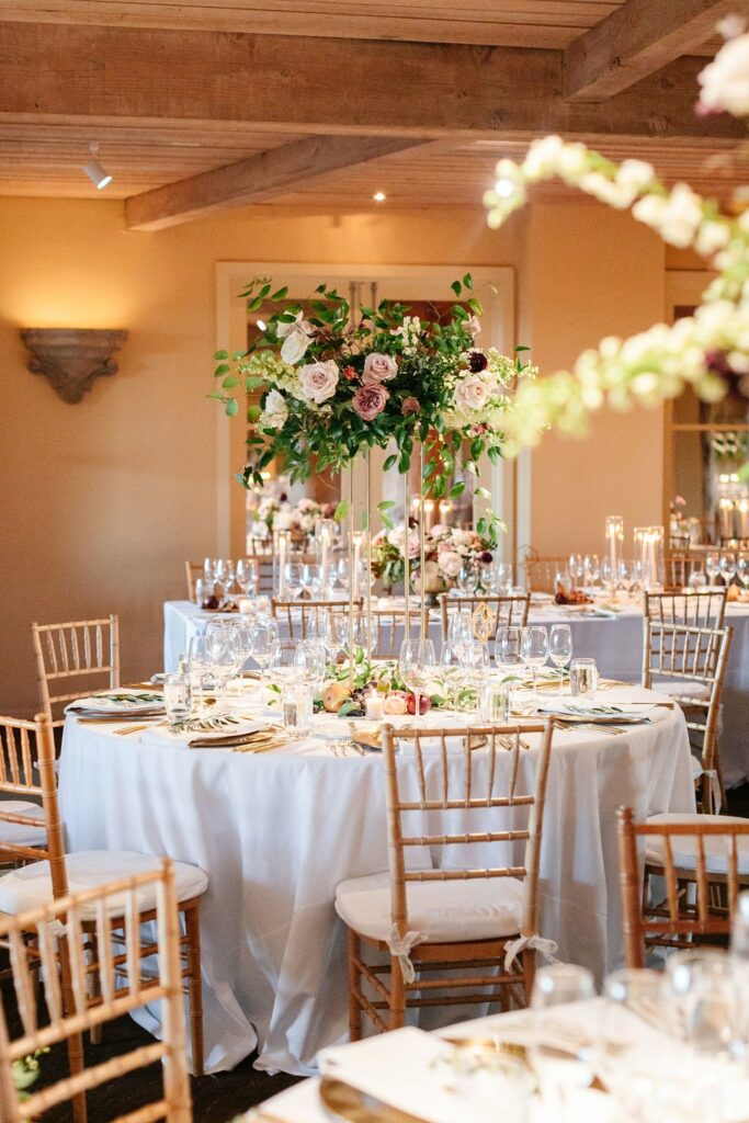 An elegantly set dining table features tall floral centerpieces, gold-rimmed glassware, and white tablecloths, surrounded by wooden chairs in a warmly lit room.