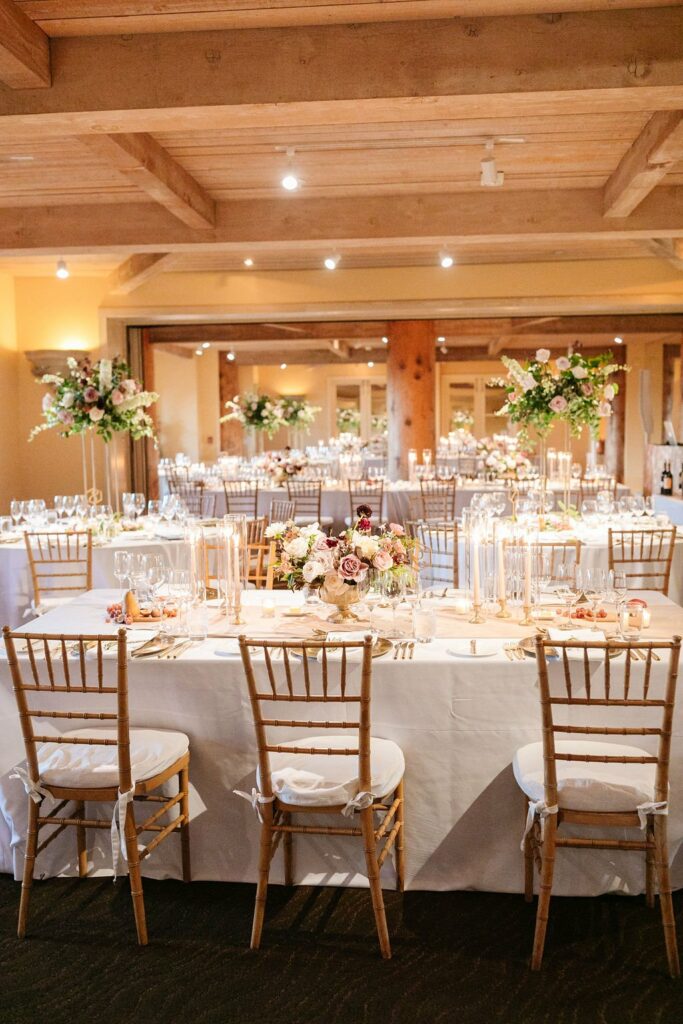 Elegant indoor wedding reception setup with white tablecloths, candles, and floral centerpieces. The room features wooden beams and warm lighting, with rows of tables and chairs neatly arranged.