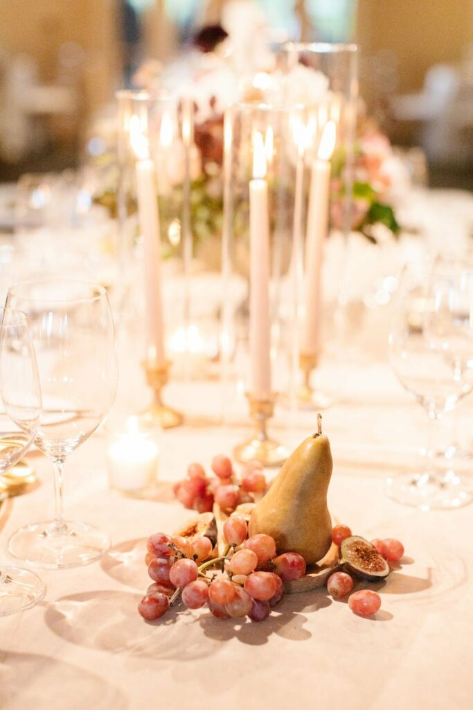 A table centerpiece featuring lit candles, grapes, a pear, and figs arranged on a white tablecloth surrounded by empty wine glasses.