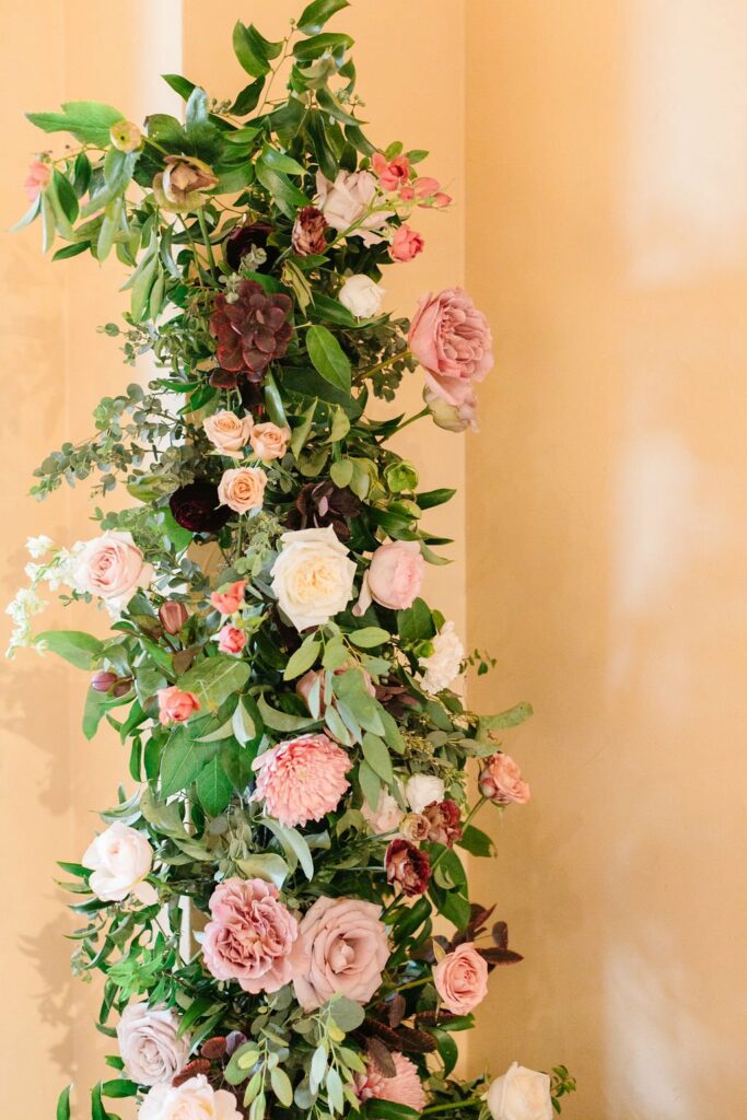 A tall floral arrangement with various flowers such as pink roses, white roses, and green foliage against a light beige wall.