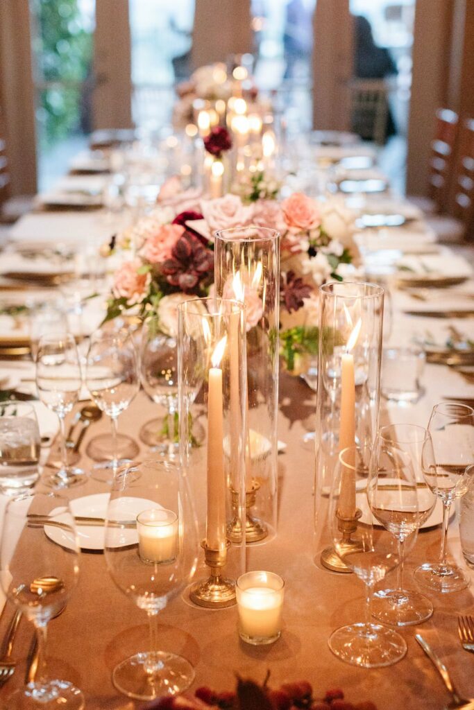 Elegant dining table set for a formal event with tall lit candles, floral centerpieces, glassware, plates, and cutlery. The background shows an indoor setting with natural light coming through windows.