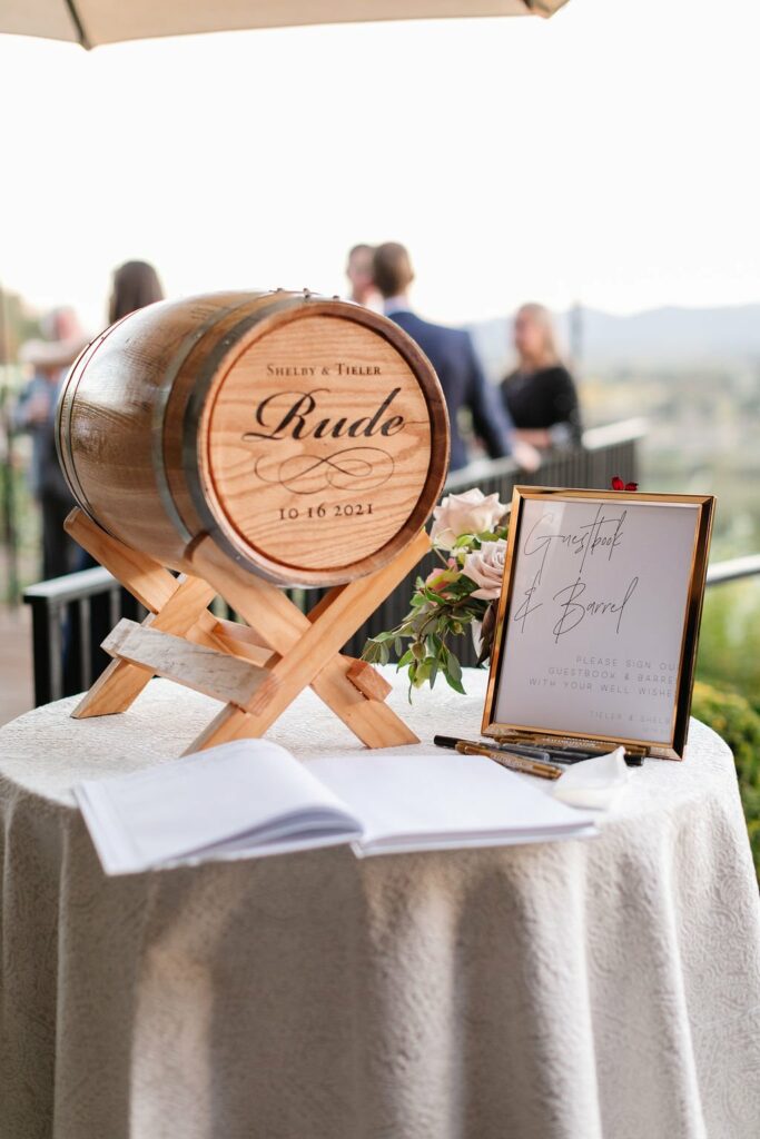 A wooden barrel marked "Shelby & Tyler, 10-16-2021" on a stand, resting on a table with a guestbook, pen, and a sign instructing guests to sign the barrel. Some people are visible in the background.