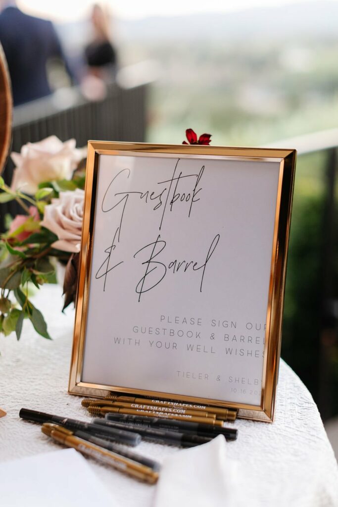 A framed sign on a table reads, "Guestbook & Barrel. Please sign our guestbook & barrel with your well wishes. Tieler & Shelby 10.16.20." Pens and a decorative arrangement are placed beside it.