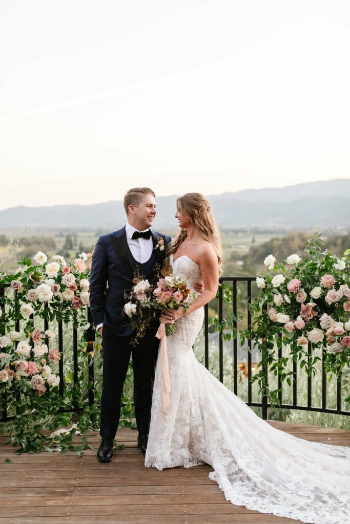 A bride and groom stand on a wooden deck with scenic rolling hills in the background. The bride is in a white lace gown, and the groom is in a black tuxedo. Both are holding a bouquet of flowers.