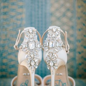 Close-up of the back of a pair of white high-heeled shoes adorned with intricate jewel embellishments, set against a patterned background.