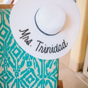 A wide-brimmed white hat with the text "Mrs. Trinidad" embroidered on it rests on a turquoise-patterned chair.