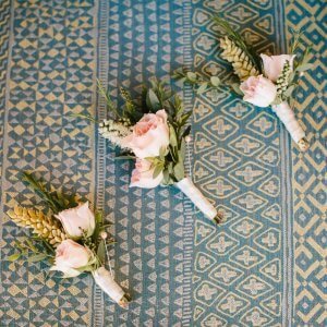 Three boutonnieres with pink roses and green foliage lying on a patterned blue-green fabric.