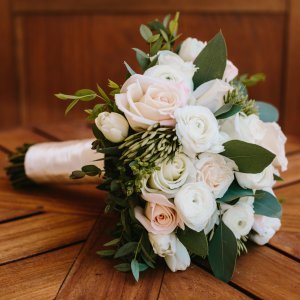 A bouquet of white and pale pink roses with green leaves, wrapped with a white ribbon, is placed on a wooden surface.