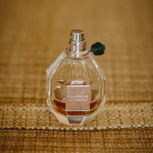 A hexagonal perfume bottle labeled "Flowerbomb Viktor & Rolf" with a green adornment on the spray nozzle, sits on a woven, tan surface.