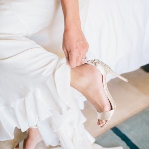 A seated person in a white dress is adjusting a high-heeled sandal on their foot.