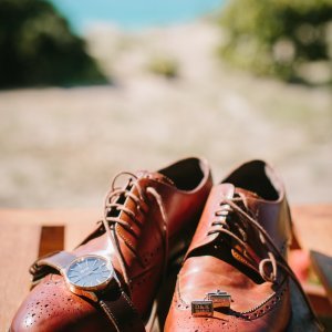 A close-up of a pair of brown leather dress shoes with a wristwatch draped over one shoe and cufflinks placed beside them, set against an outdoor background.