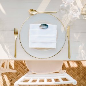 A white table setting with a gold-rimmed plate, white napkin, name card, gold utensils, and two clear glasses.