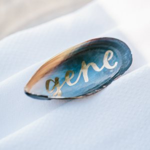 Close-up of an open mussel shell with the name "Gene" written in gold cursive, placed on a white textured fabric.