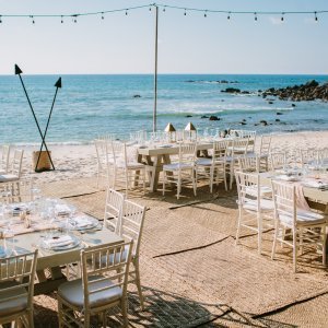 Beachfront dining setup with white wooden chairs and rectangular tables on a sandy shore, decorated with string lights and triangular decor, overlooking the ocean, under a clear blue sky.