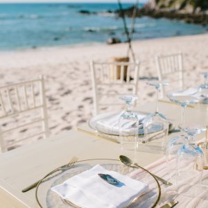 Beachside table setting with clear plates, white napkins, and glassware, positioned on a sandy beach with the ocean in the background.