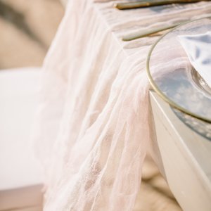 Close-up of a table draped with a light pink, semi-transparent fabric. Two metallic cutlery pieces rest beside the glass plate with a folded white napkin. White and beige shades dominate the background.