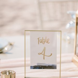 A sign displaying "Table 4" in gold text stands on a light-colored table adorned with gold candle holders and soft decor elements.